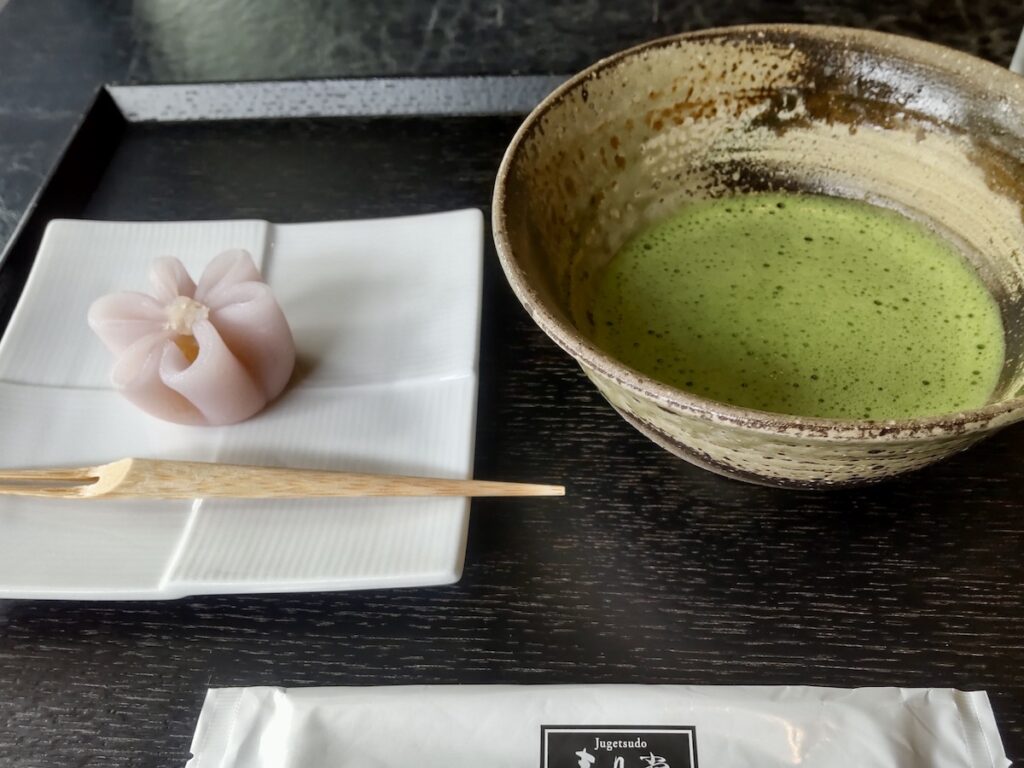 A cup of frothy green matcha tea served with a pink wagashi shaped like a clematis flower
