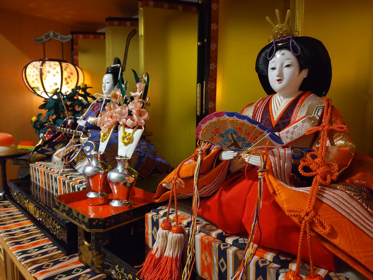 Close-up of the female Hina doll in a vibrant entryway display with gold folding screen, lanterns, and flowers
