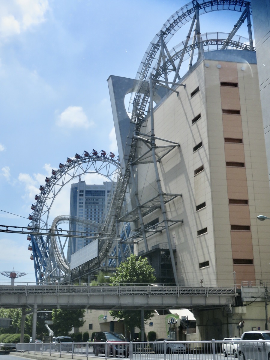 An outdoor view of Tokyo Dome City featuring a blue roller coaster track weaving through buildings in a dense urban setting