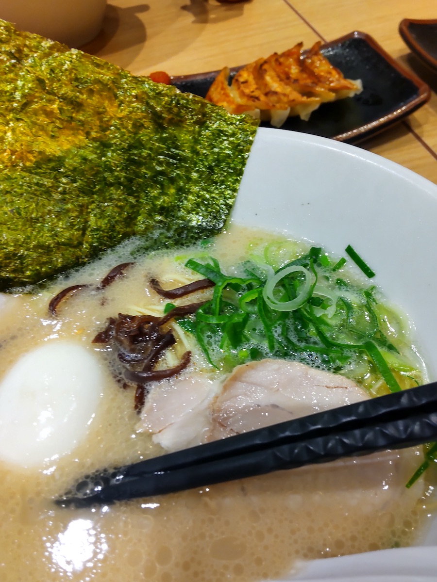 A bowl of creamy white soup with green onions, chashu, chopsticks, and gyoza
