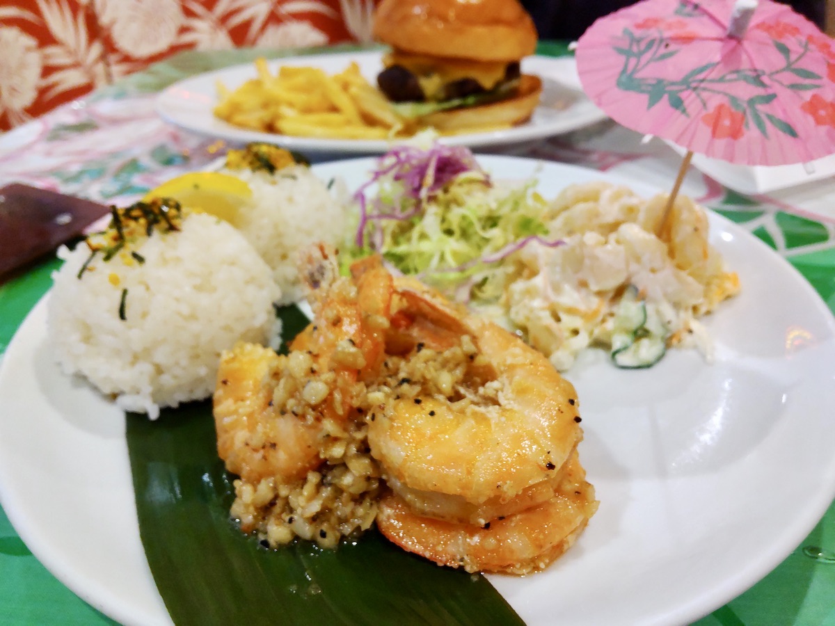 Garlic shrimp plate with a small decorative umbrella and a hamburger in the background