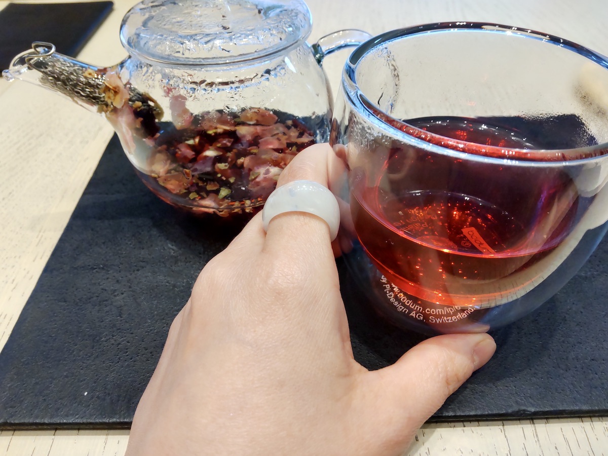 A hand touching a teapot filled with red petals and a cup of red tea