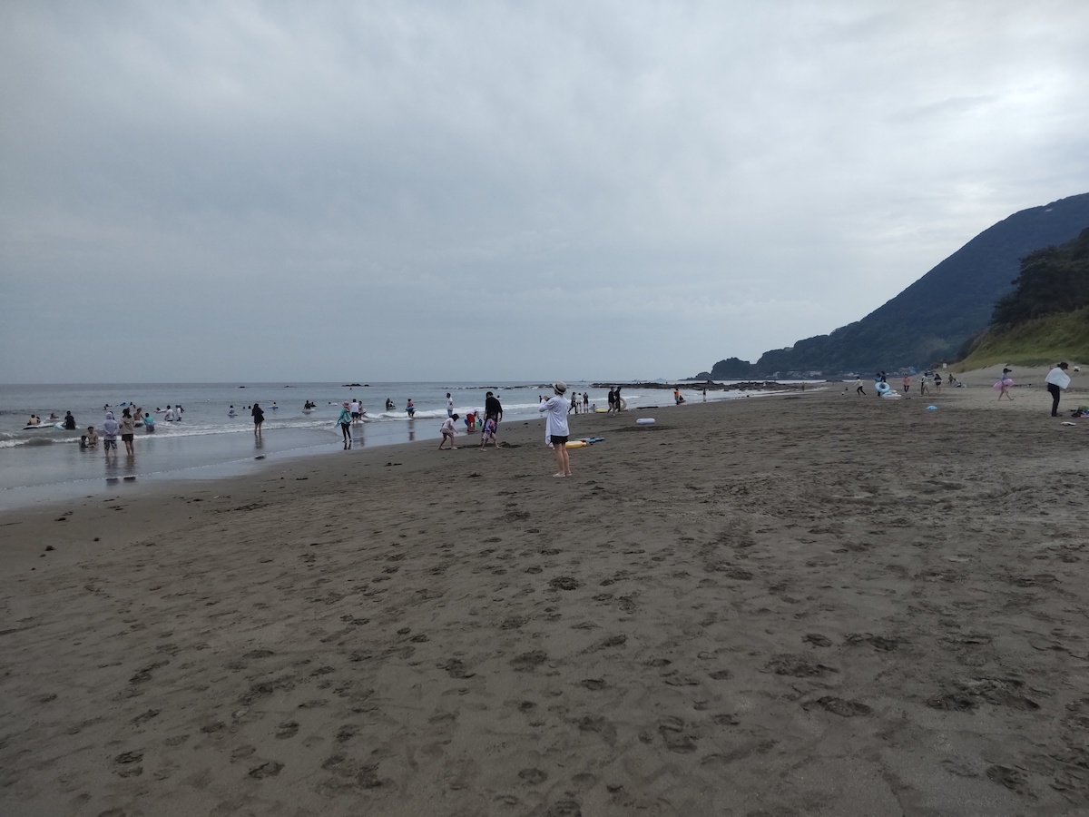 A cloudy beach and sea with people playing, and mountains in the background