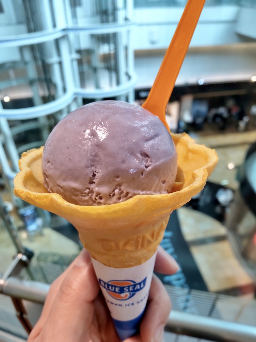 A hand holding purple Blue Seal ice cream in a cone inside an airport terminal