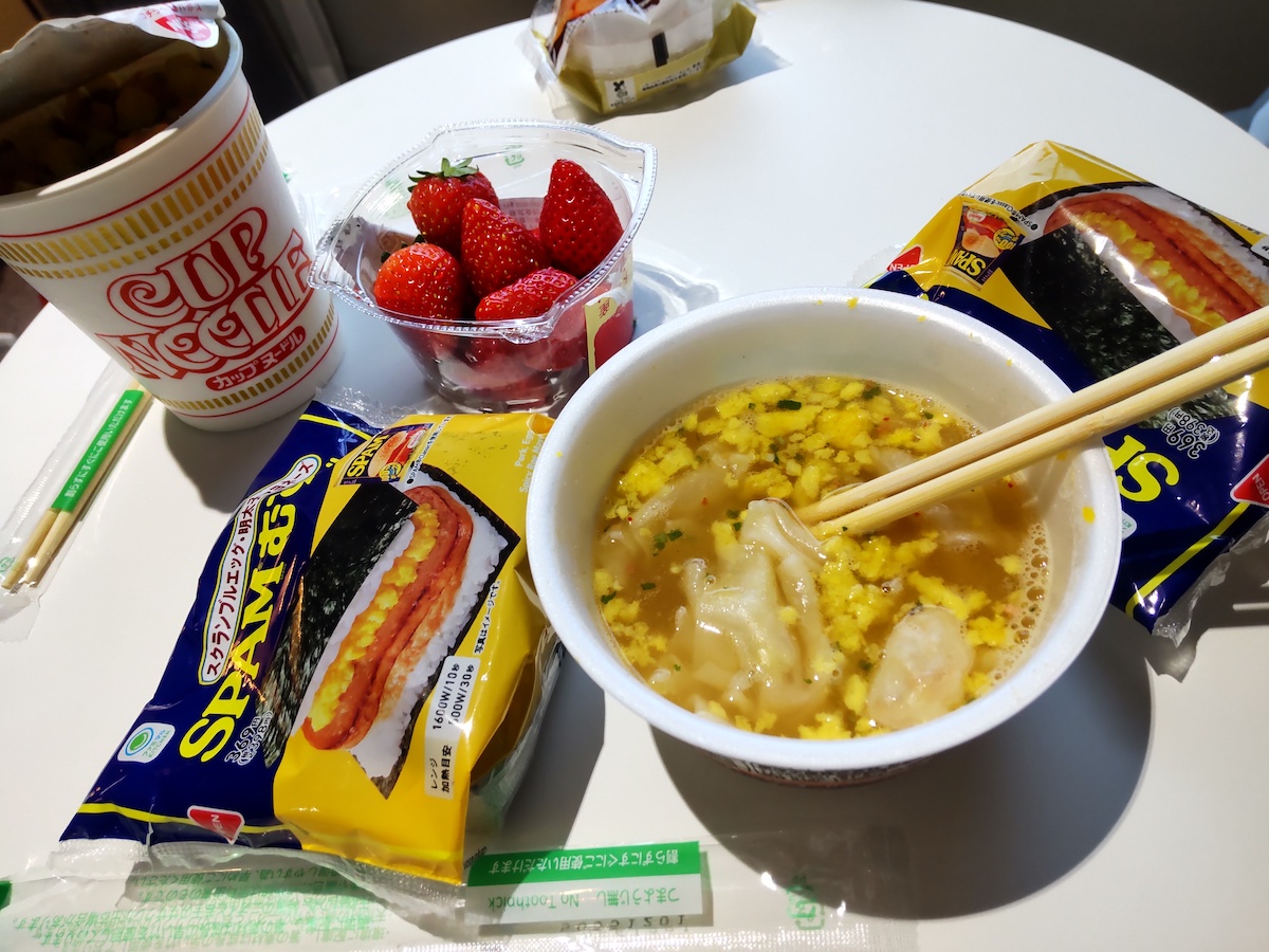 A spread of Japanese convenience store food, including cup noodles, Spam musubi, and strawberries