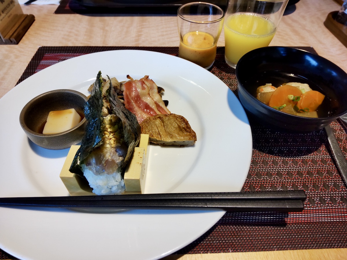A Japanese-style breakfast featuring an open onigiri, side dishes, soup, and a beverage