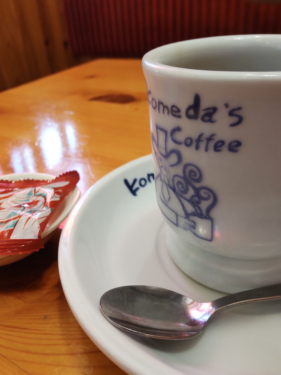Komeda Coffee mug on saucer served with packaged nuts on wooden table