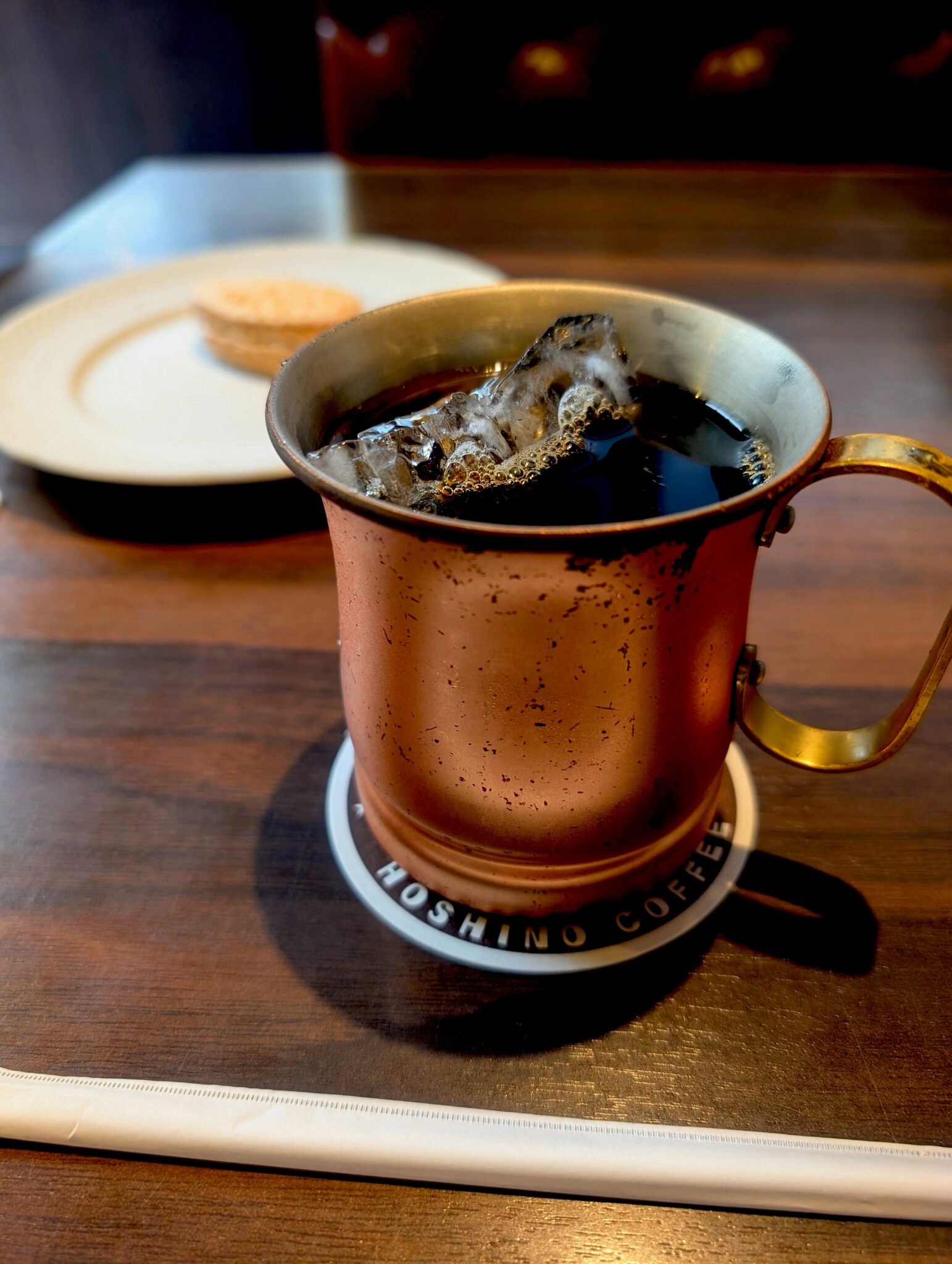 Iced coffee in copper mug on Hoshino Coffee coaster with dessert behind